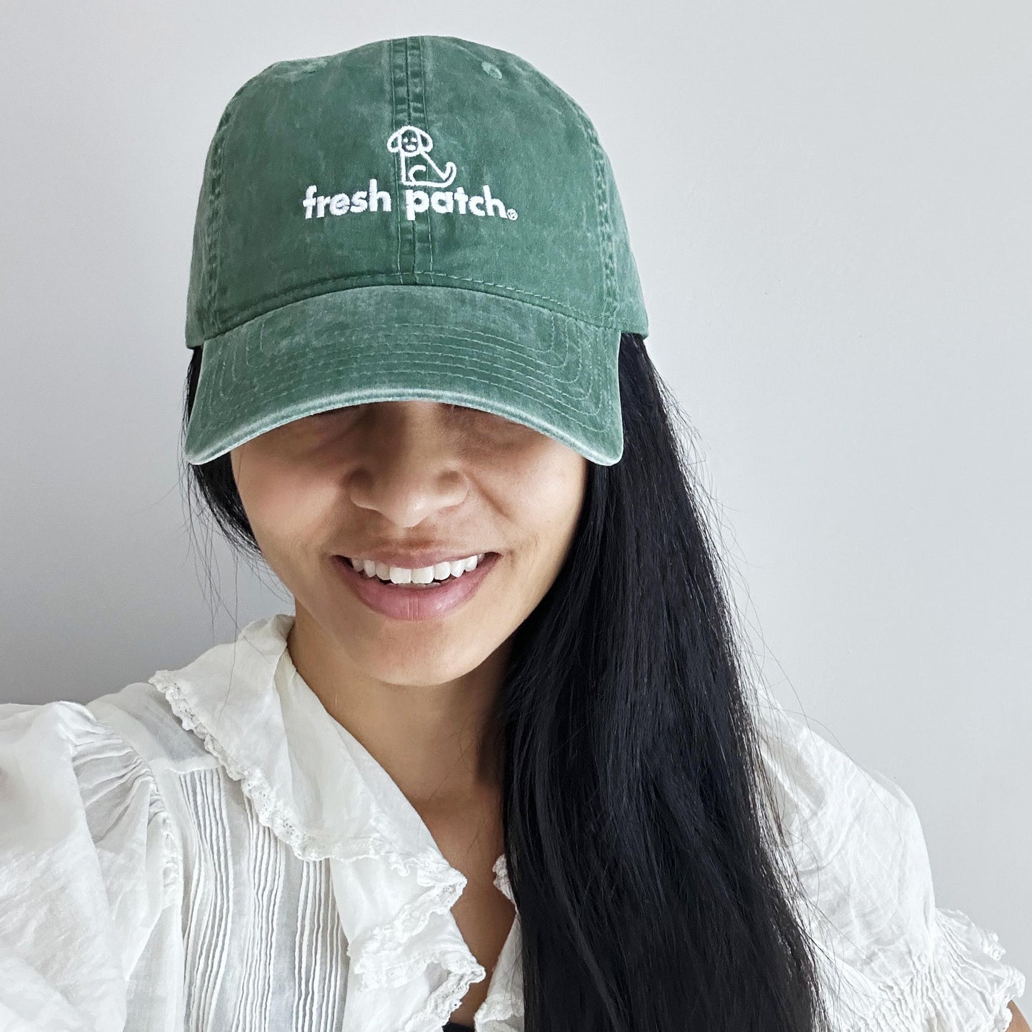 Girl with long black hair wearing greenish baseball cap with 'fresh patch' branding, facing forward