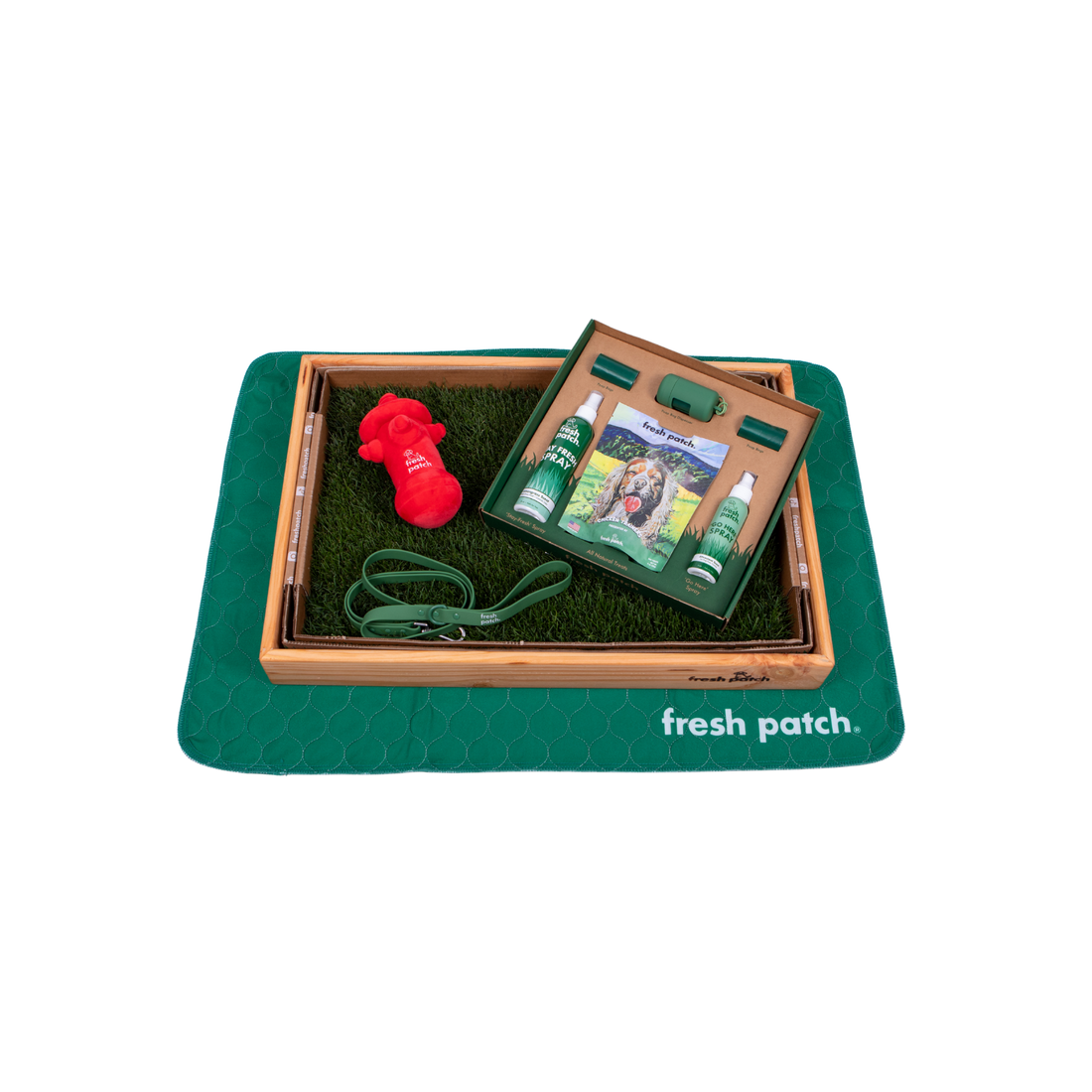 A top-down view of a Fresh Patch wooden grass tray on a green mat, holding a leash, red fire hydrant toy, and an open accessories kit.