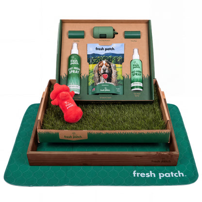 Fresh Patch potty training kit with a real grass tray in a dark wooden frame, a plush hydrant toy, two sprays, dog treats, poop bags and a green pad.